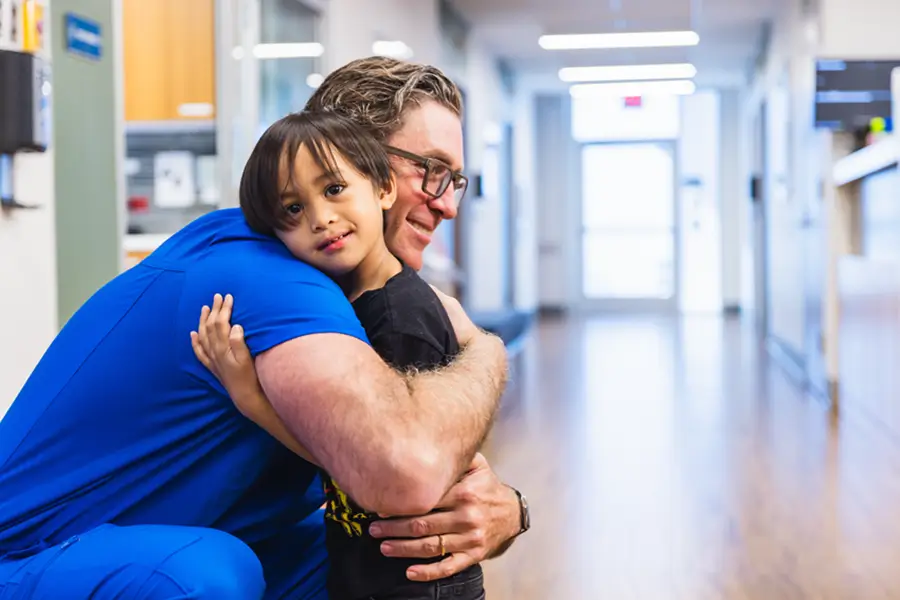 Physicians in blue scrubs hugging young child in emergency room hallway.